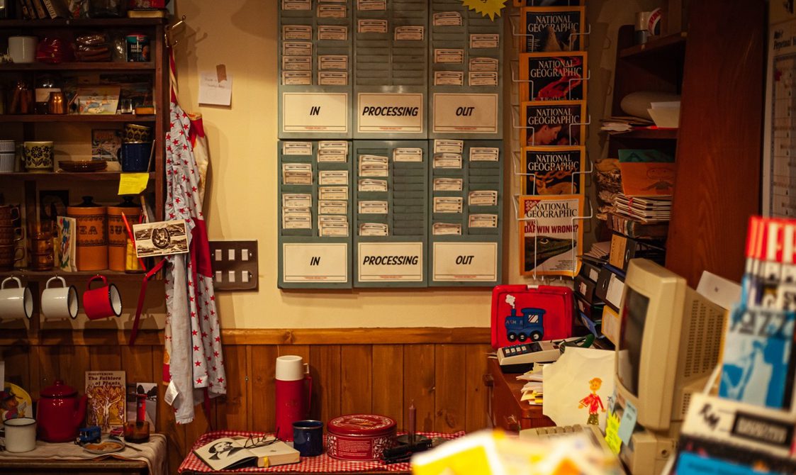 an image of inside our venue. The set design is inspired by a bric-a-brac shop, with lots of objects, papers and shelves. There is a faded turquoise board with signs saying 'in', 'processing' and 'out'.
