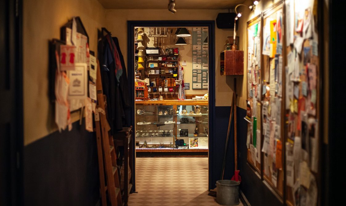 An image of the hallway leading up to the door to enter inside our venue, Punchdrunk Enrichment Stores. The hallway has a board on the right hand side with many leaflets and papers pinned up. There is a metal buckets near the doorway on the right hand side and a folded up ladder on the left hand side.
