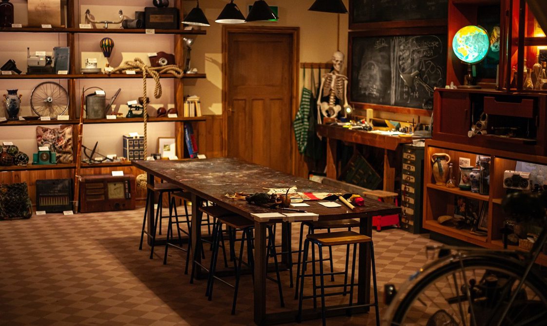 A full view inside of Punchdrunk Enrichment Stores. The main view shows a wooden table on a brown checker floor. Around the inside of the venue is shelves with lots of curious objects. In the distance is a wooden door.