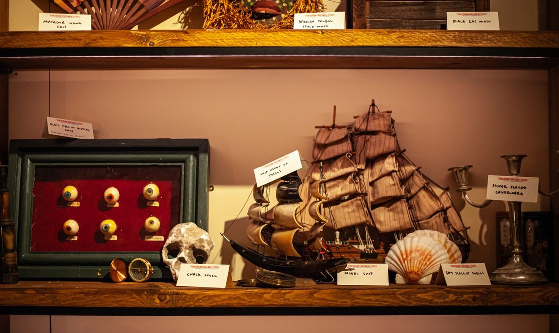 A close up image inside the venue of objects along a shelf. Objects include a ship, seashells, a frame and a fan.