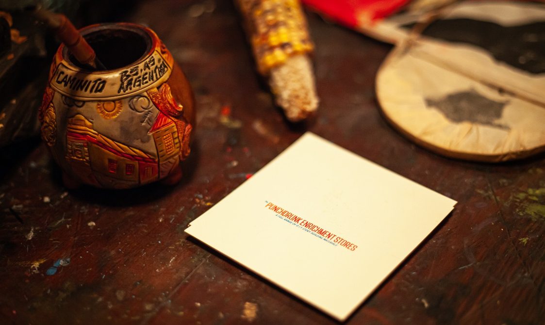 A close up image of a plain card with bold red text saying 'Punchdrunk Enrichment Stores', sitting on a wooden table. Around the card, there are decorated objects on the table.