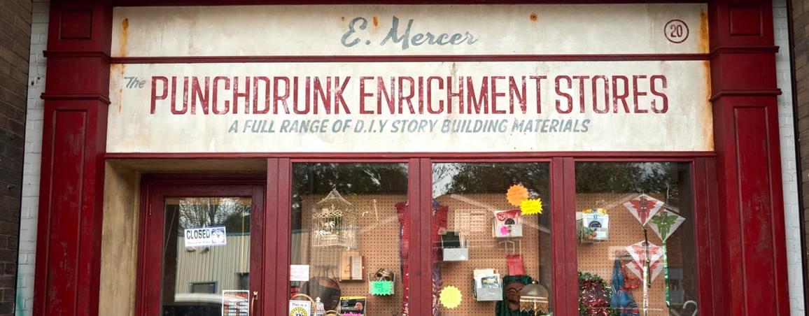 Image of the Punchdrunk Enrichment Stores shopfront. It is clad in red wood, with an aged sign that reads 'Punchdrunk Enrichment Stores' with the words 'A full range of DIY story building materials' underneath. There are three windows which show a number of items including a birdcage, a monopoly set and framed photographs. There is a door to the shop with opening times displayed.