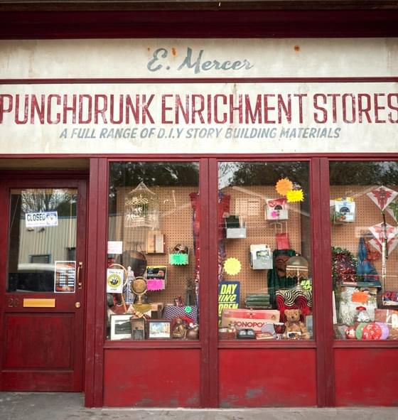Image of the Punchdrunk Enrichment Stores shopfront. It is clad in red wood, with an aged sign that reads 'Punchdrunk Enrichment Stores' with the words 'A full range of DIY story building materials' underneath. There are three windows which show a number of items including a birdcage, a monopoly set and framed photographs. There is a door to the shop with opening times displayed.
