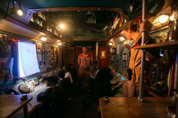 Children and two performers wearing jumpsuits are in what looks like a spaceship control room with lots of dials and switches on the walls.