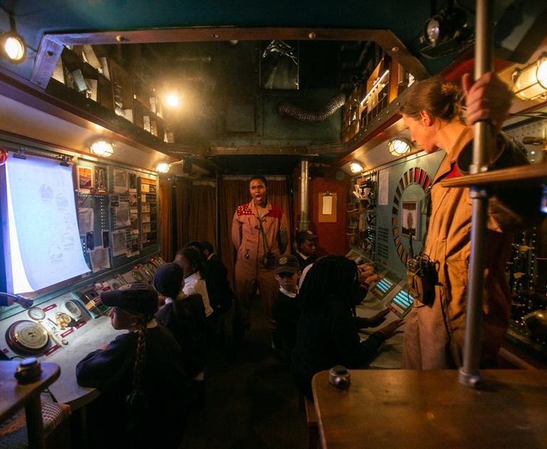 Children and two performers wearing jumpsuits are in what looks like a spaceship control room with lots of dials and switches on the walls.