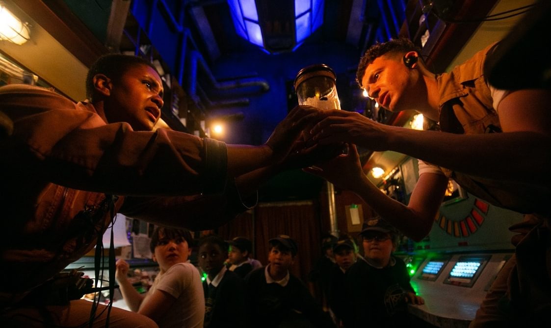 The camera is looking up at two performers wearing boiler suits, they are both holding a jar with something inside. Their facial expressions both look concerned. They are inside a bus which looks like the control room of a spaceship. Above them an atrium is glowing blue. Behind them you can see schoolchildren, seated and staring on with amazement.