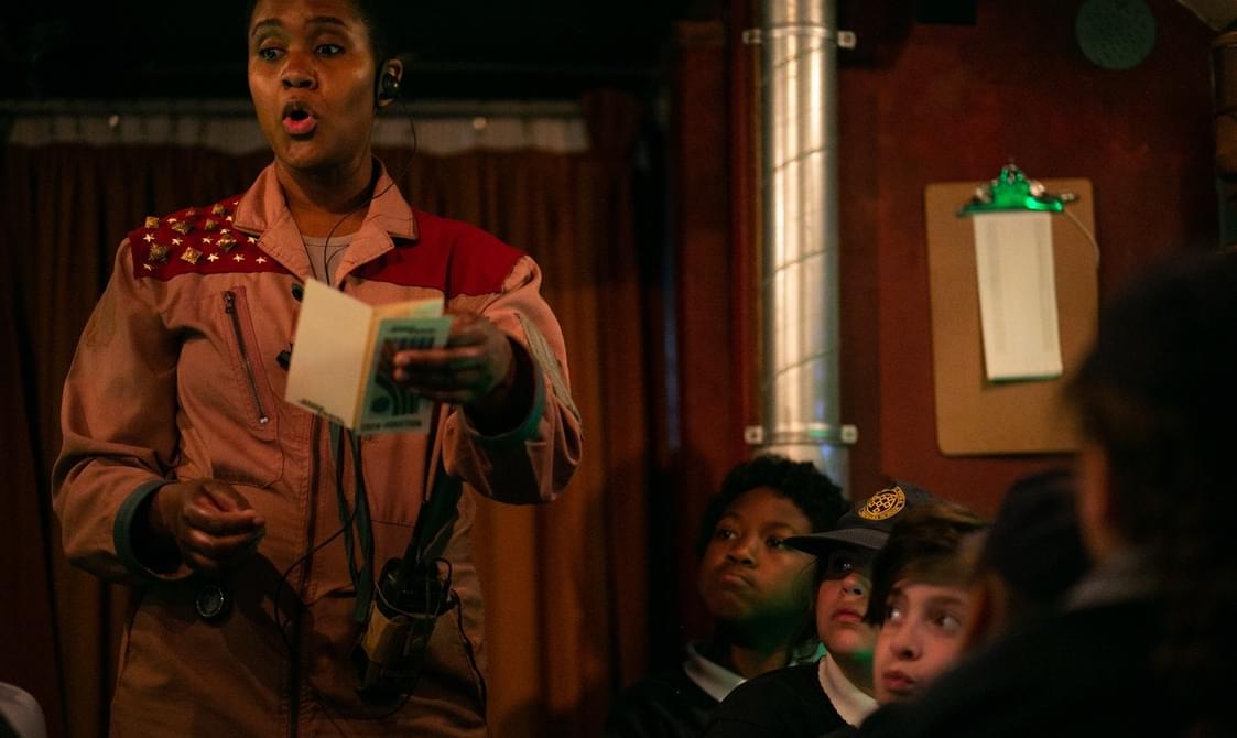 A performer wearing a peach coloured boiler suit with studs on one shoulder and wearing an earpiece is holding up a small booklet whilst speaking. Three schoolchildren are seated to her right and and look on.