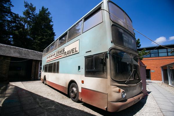 A dusty old double decker bus parked outside in the sunshine. 'Ermis Travel' is written on the side of the bus. It is a light blue at the top with white and red at the bottom.
