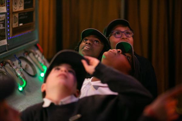 Four school children are sat inside what looks like a space ship control room. They are all looking up, transfixed.