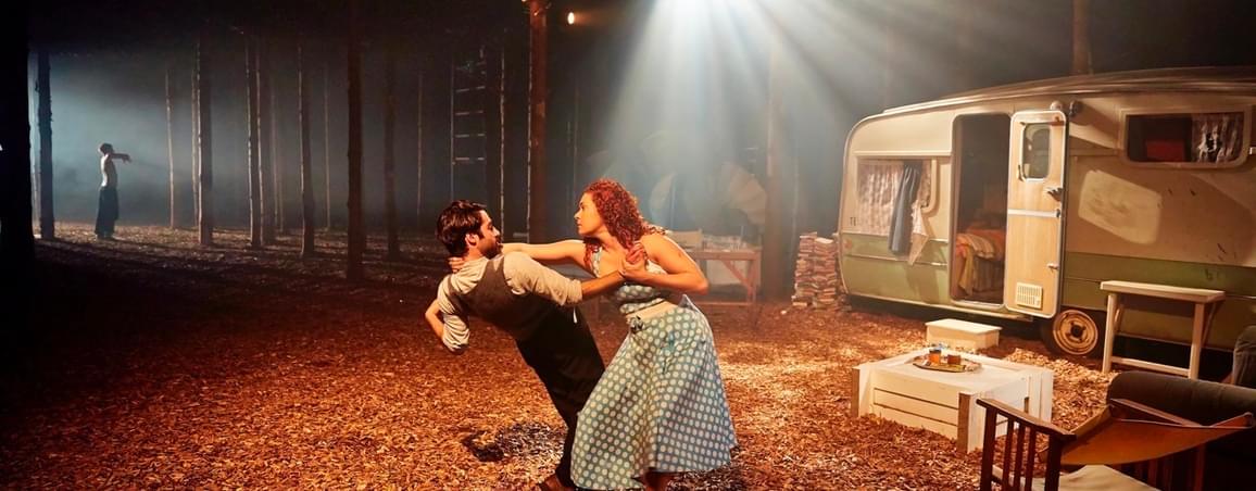 Two performers dance together in a woodland forest. There is a caravan behind them.