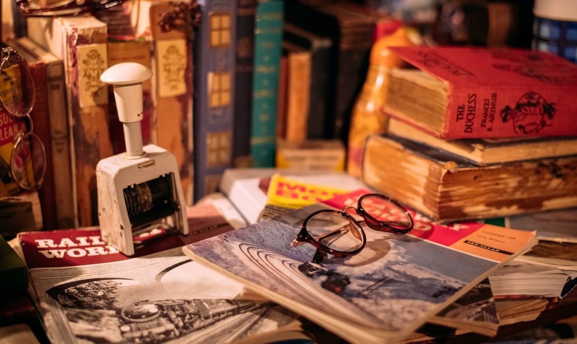 An image of a pile of books and magazines on the table with a pair of glasses in the foreground.
