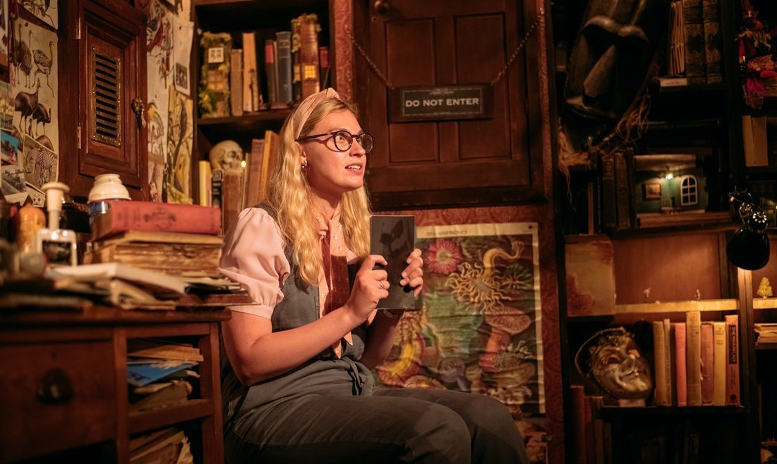 In a dimly lit room, a woman is clasping tightly a closed book in front of her chest with both hands. She is wearing glasses and a pink hairband. She is wearing grey dungarees with a pastel pink, puffy sleeve blouse. She is sitting on a chair and surrounded by books, items and shelves.