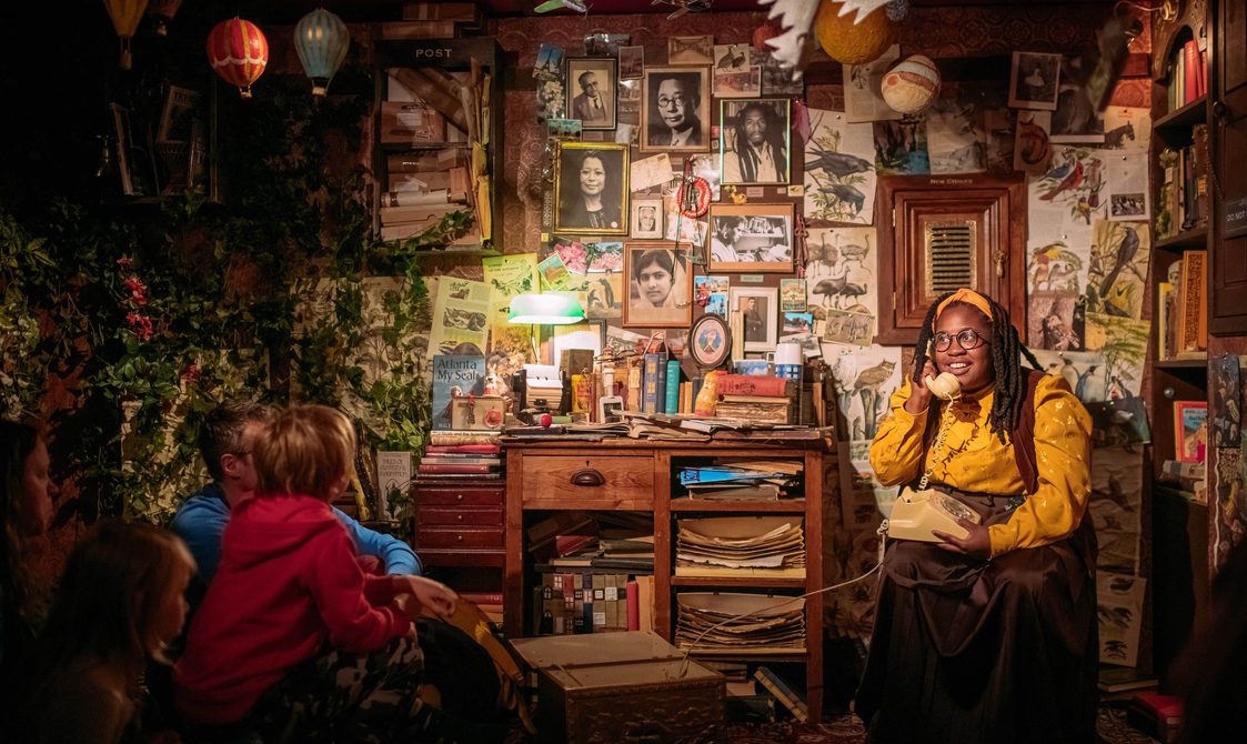 In a dimly lit room full of books, a library guardian wearing a yellow shirt and headband answers the phone.