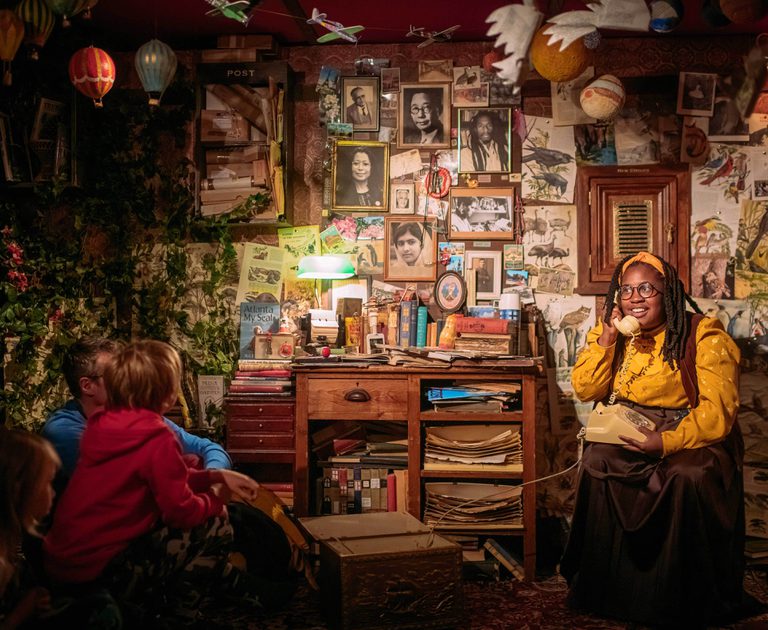 In a dimly lit room full of books, a library guardian wearing a yellow shirt and headband answers the phone.