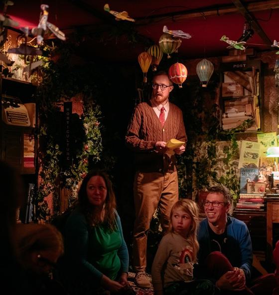 In a dimly lit room surrounded by books, a library guardian wearing a brown cardigan and glasses stands over a group of children and their families. There are tiny planes and hot air balloons around his head.