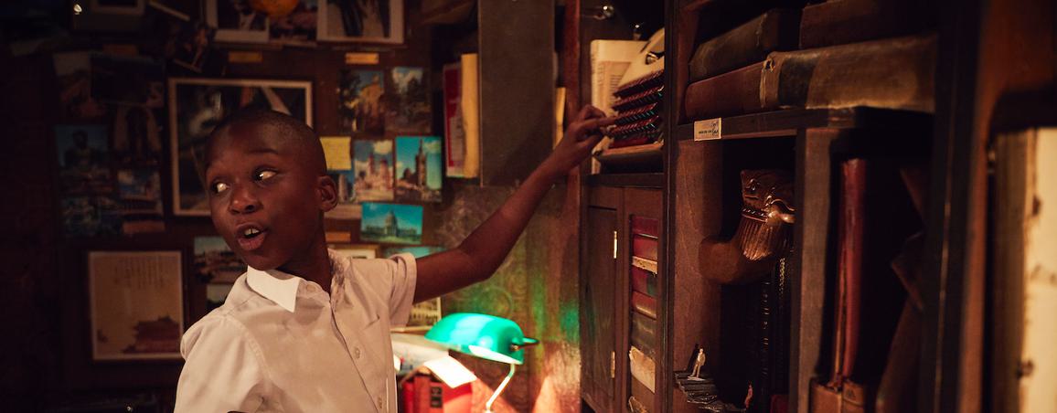 A young boy smiling and pointing at at a shelf of books in a dimly lit library.