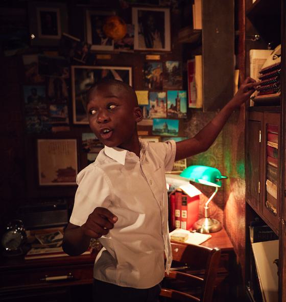 A young boy smiling and pointing at at a shelf of books in a dimly lit library.