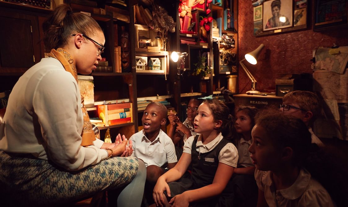 Image of a woman sat on a chair reading to school children who look up to her in amazement. She is telling them a story. They are sat in a small room with lots of books and pictures crammed onto shelves. It is a dark room with red walls and low lighting.