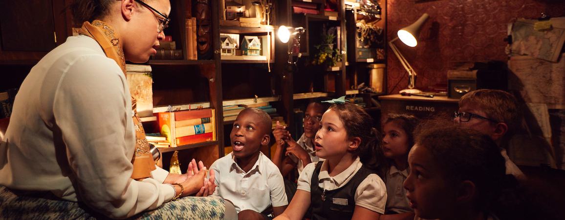 Image of a woman sat on a chair reading to school children who look up to her in amazement. She is telling them a story. They are sat in a small room with lots of books and pictures crammed onto shelves. It is a dark room with red walls and low lighting.