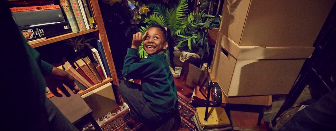 In a room full of books, plants and boxes, a young girl is kneeling down, looking back and smiling.