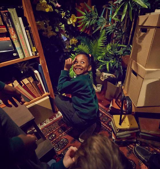 In a room full of books, plants and boxes, a young girl is kneeling down, looking back and smiling.