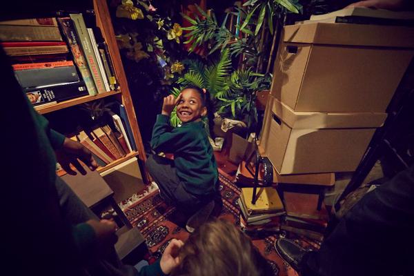 In a room full of books, plants and boxes, a young girl is kneeling down, looking back and smiling.