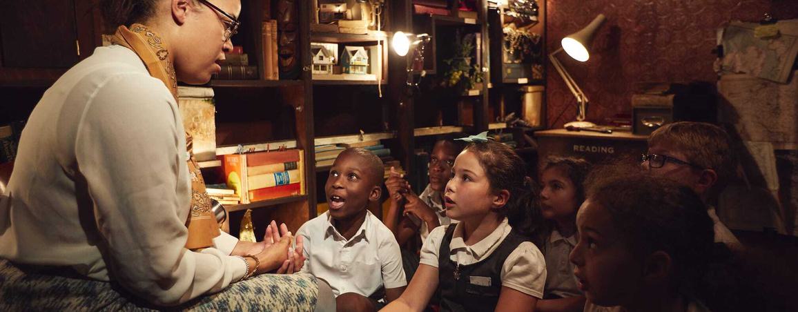 In a dimly lit room surrounded by books, a performer is sat on a chair in front of a group of children sat on the floor