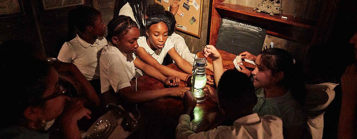 A group of children in a dimly lit room stand around a table and hold a green lantern.