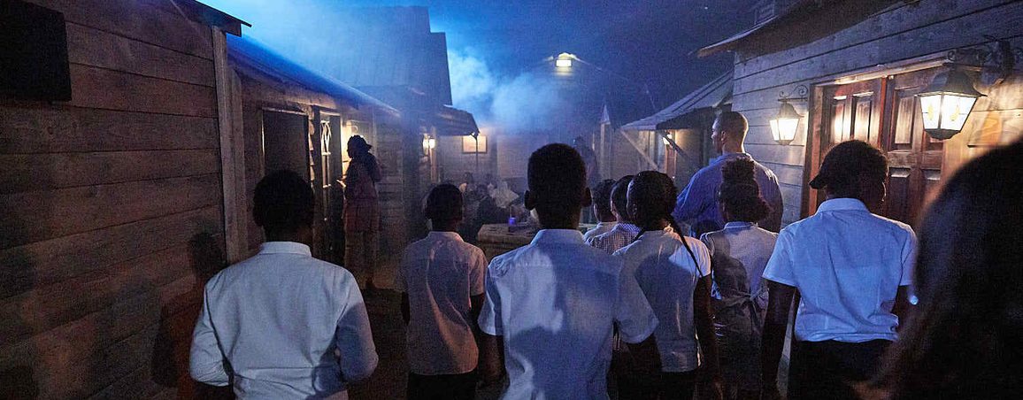 In a dimly lit environment, a group of school pupils look into the distance. There is smoke, haze and blue ellipsoidal spotlights in the village of Fallow Cross.