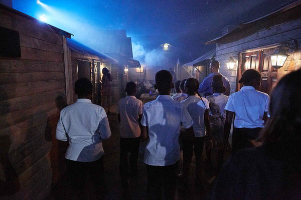 In a dimly lit environment, a group of school pupils look into the distance. There is smoke, haze and blue ellipsoidal spotlights in the village of Fallow Cross.