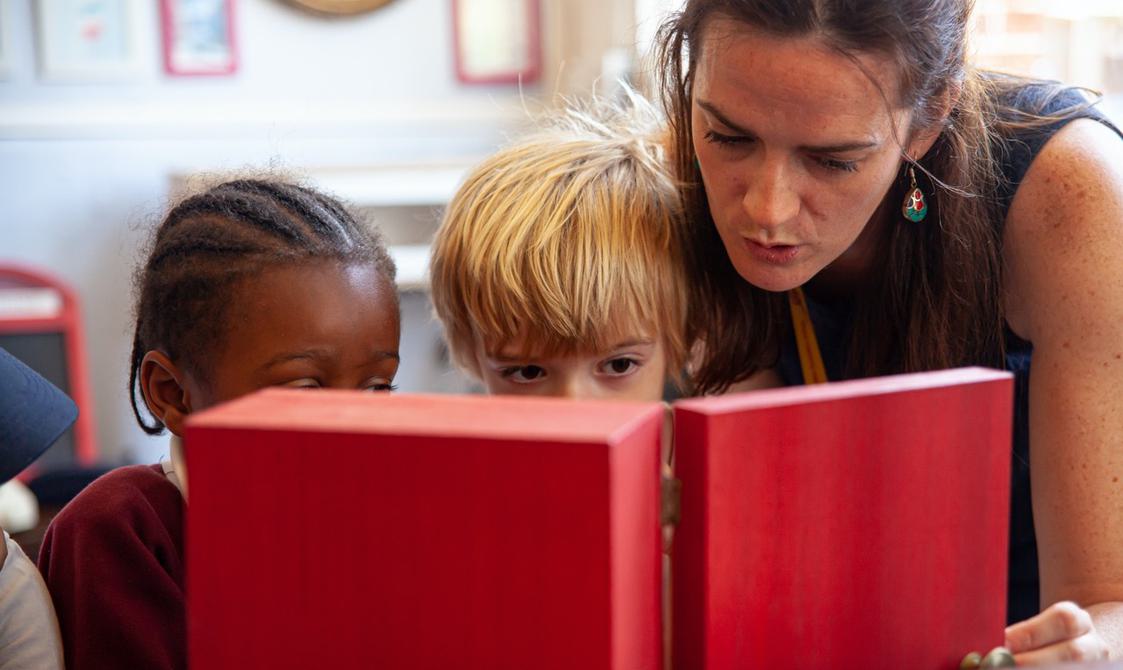 A teacher and two young children look inside a red cupboard.