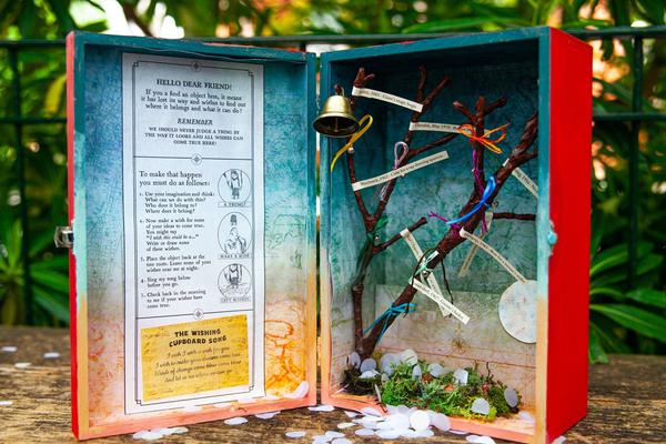 'The Wishing Cupboard' is placed open on a garden wall. It is a small red box, inside is a tree with ribbons and a bell, a map, and some paper instructions.