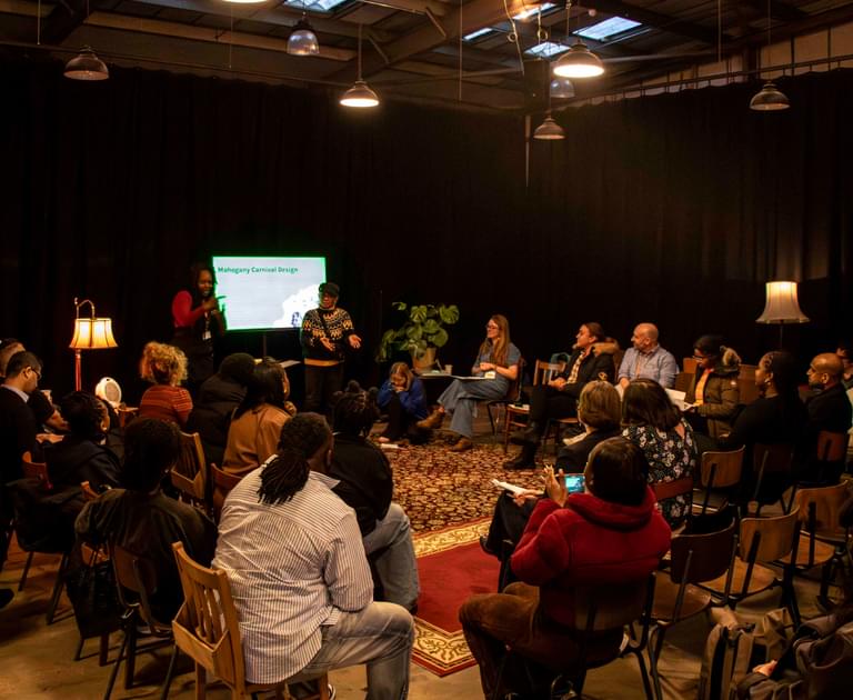 Two women standing by a screen, presenting to a group of people seated in a half circle in front of them